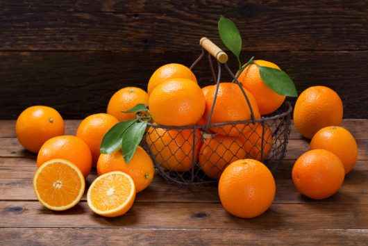 A wire basket filled with Citrus Orange Tree 'Seville' oranges, some with leaves still attached. Whole oranges are scattered around, and one orange is cut in half, revealing the vibrant inside. The background features a wooden surface and wall, reminiscent of a rustic Citrus Orange Tree orchard.