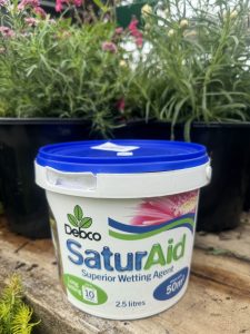 A bucket of debco saturaid wetting agent placed in front of blooming pink flowers, suggestive of use in gardening.
