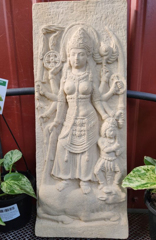 Stone Iridami Relief Dewi Sri White 20x3x50cm sculpture depicting Dewi Sri, a multi-armed deity, standing with a small figure and a lion at their feet. Surrounded by various objects, the deity is adorned in detailed attire.