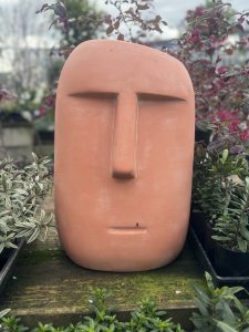 A Moai Statue Terracotta 30x29x46cm stands amidst potted plants displaying various shades of green and purple.