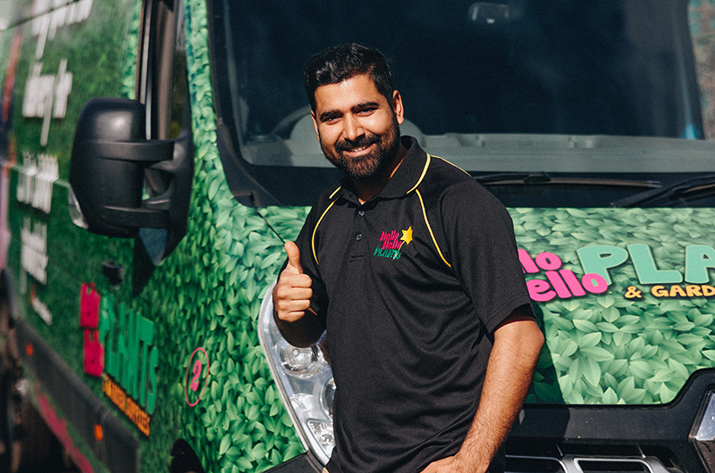A man with a beard gives a thumbs up while standing in front of a green vehicle with a leafy design, advertising bare rooted plants for sale. He is wearing a black polo shirt with a company logo.