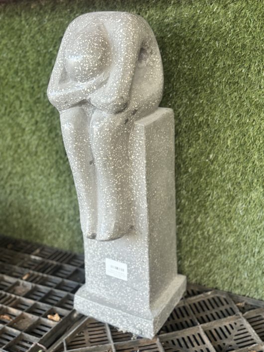 A stone statue of a person sitting with their head resting on their knees, mounted on a rectangular pedestal, stands in front of an Iridami Resting Man Grey Terrazzo 16x20x60cm textured background.