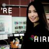 A woman stands in front of two computer monitors with a website displayed, next to the words "We're Hiring." She is wearing a black top and smiling confidently at the camera, embodying our enthusiastic hiring spirit.