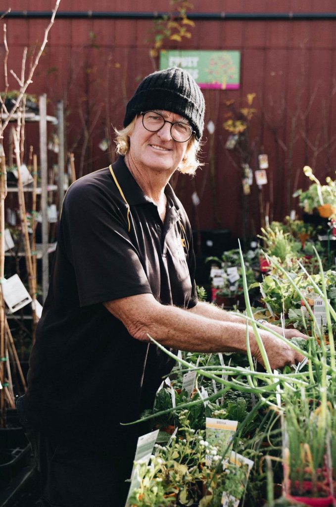 A person wearing a black beanie and glasses stands in a garden center among bare rooted plants for sale, smiling at the camera.