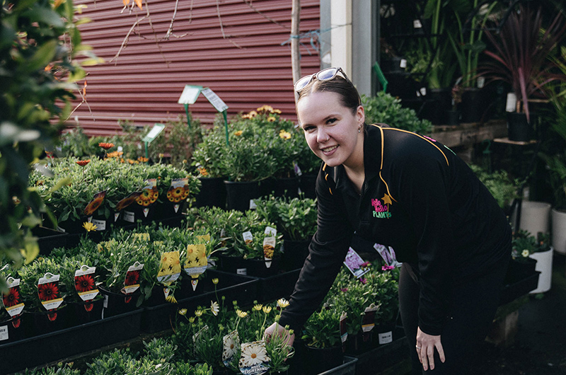 A person in a black shirt is picking plants in a garden center, with various potted flowers and bare rooted plants for sale arranged on shelves around them.