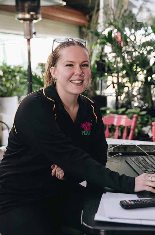 Woman smiling, seated at a table with a laptop, remote, and paper, wearing a black top with "Hello Hot Plants" logo. Bare rooted plants for sale are visible in the background among the chairs and greenery.