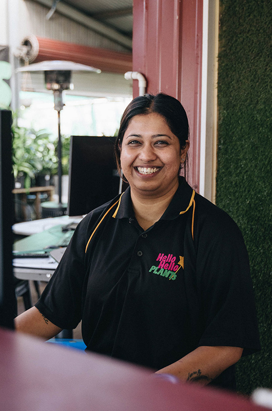 Smiling woman in a black polo shirt with "Hello Hello Plants" logo stands indoors, surrounded by various equipment and a green wall, showcasing bare rooted plants for sale.