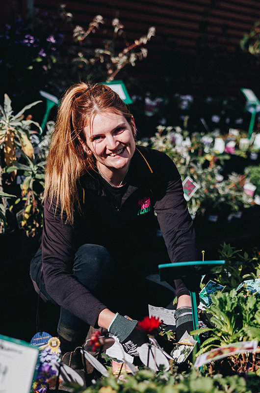A person crouches while tending to bare rooted plants for sale in a garden center, smiling at the camera. They are wearing gardening gloves and a dark uniform.