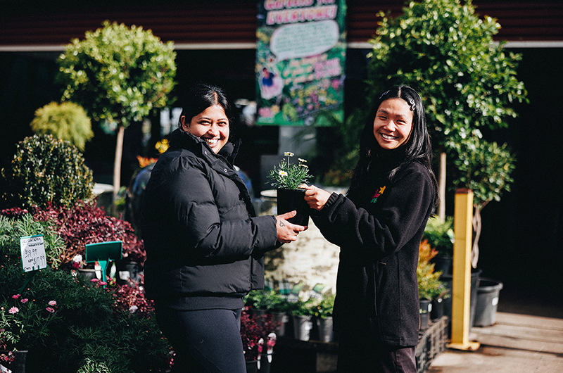 Two women smiling and holding a potted plant together in an outdoor plant nursery, where bare rooted plants for sale are displayed among the rich greenery in the background. Hello Hello Staff Photo, Employee