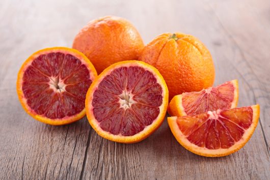 Two whole and two halved blood oranges, displaying vibrant red flesh, rest on a wooden surface. These fruits highlight the rich hue and succulent nature of the prized Citrus Orange Tree 'Blood Orange' Dwarf from a 10" pot.