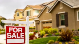 A "For Sale" sign in front of a suburban house with surrounding garden and neighboring houses in the background.