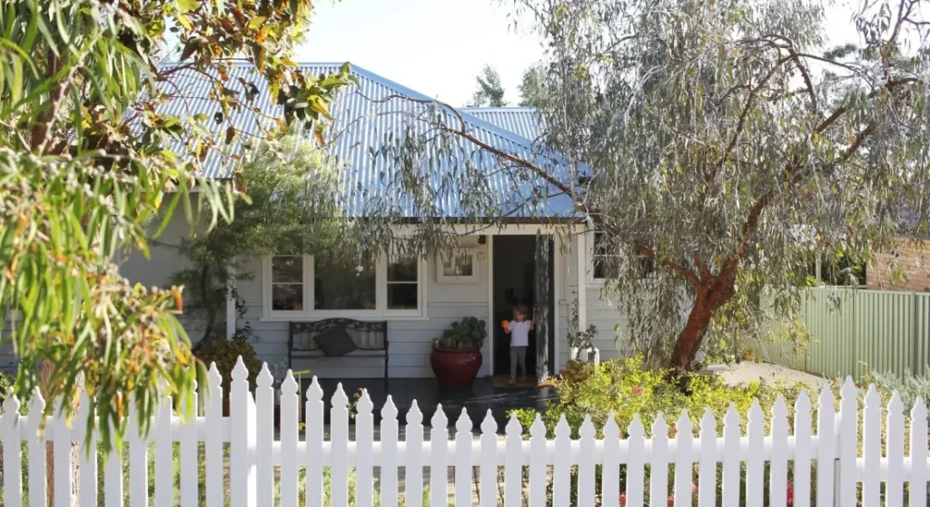 A quaint house with light grey siding, a blue metal roof, and a white picket fence features thoughtful garden design. A child and an adult are standing in the open doorway amidst greenery and trees.