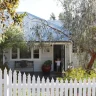 A quaint house with light grey siding, a blue metal roof, and a white picket fence features thoughtful garden design. A child and an adult are standing in the open doorway amidst greenery and trees.