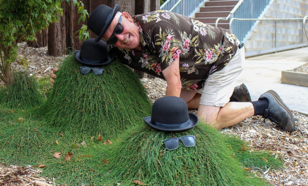 A man poses with two bush sculptures dressed with bowler hats and sunglasses, kneeling on the ground and smiling, showcasing his unique garden design.