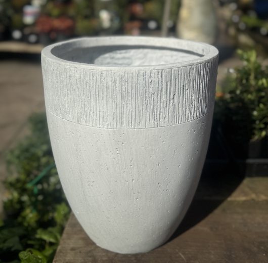 A GeoLite Tall Egg White Wash planter, sized 29x34cm and featuring a slightly tapered cylindrical shape in a large, light gray design, is showcased on a wooden surface. The textured planter stands tall with plants and other garden items visible in the blurred background.