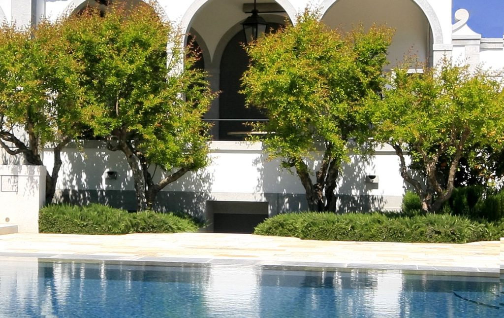 A white building with arched doorways and windows, partially obscured by trees, stands elegantly amidst thoughtful garden design, viewed from across a blue swimming pool.