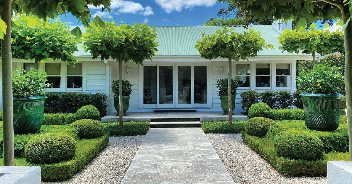 A single-story white house with a green roof and symmetrical landscaping featuring trimmed hedges, topiary, potted plants, and a stone path leading to the entrance showcases expert garden design.