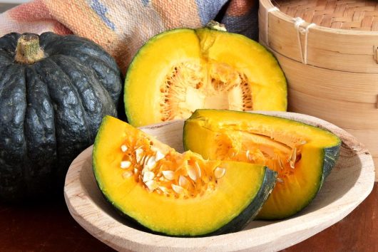 A cut Pumpkin 'Japanese' with bright yellow-orange flesh and seeds displayed in a wooden bowl, accompanied by an intact Pumpkin 'Japanese' 4" Pot and a wooden steamer in the background.