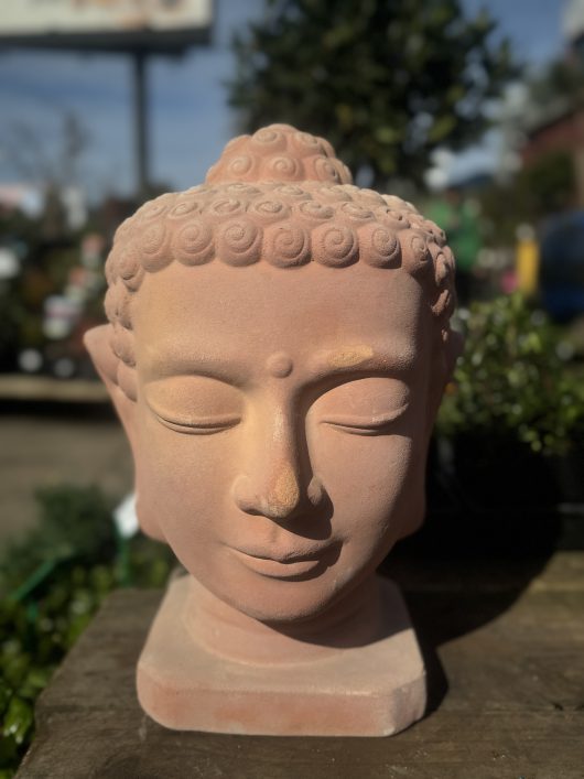 Close-up of the Terracotta Buddha Head 26x40cm sculpture, featuring a peacefully closed-eyed expression reminiscent of a Moai statue, set outdoors with surrounding greenery slightly out of focus.