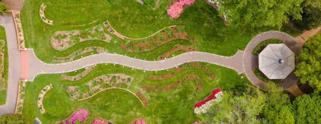 Aerial view of a landscaped garden featuring thoughtfully planned pathways, various flower beds with green and blooming plants, and a gazebo in the right corner showcasing exquisite garden design.