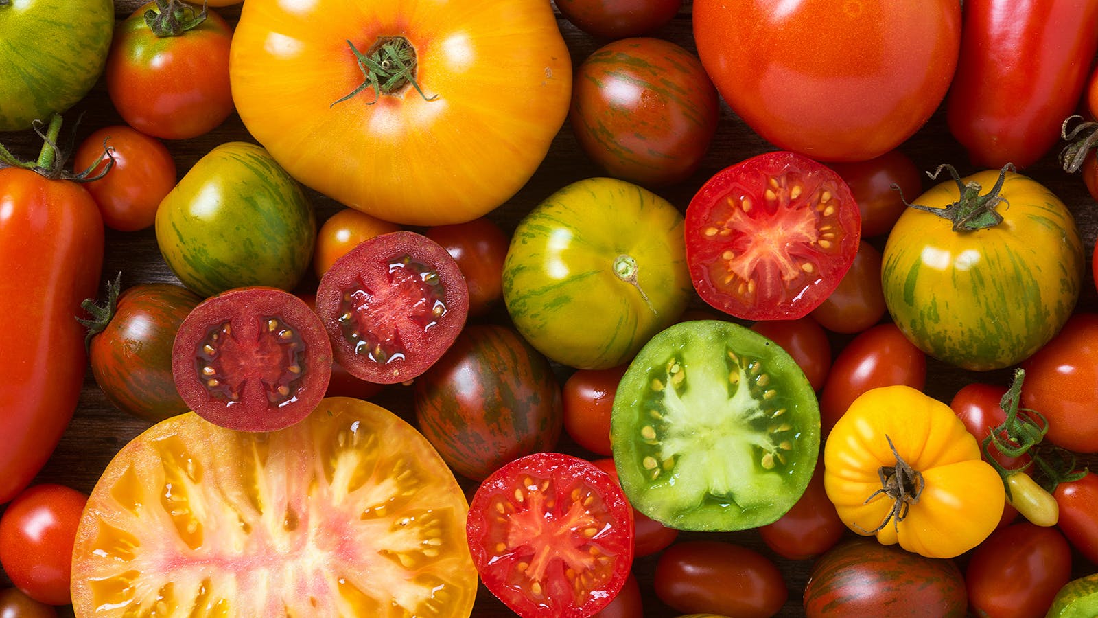 A variety of heirloom tomatoes in different shapes, sizes, and colors, including red, yellow, green, and striped, are displayed close together. Amid this vibrant display that could solve any garden design problems, some tomatoes are sliced to show their interior.