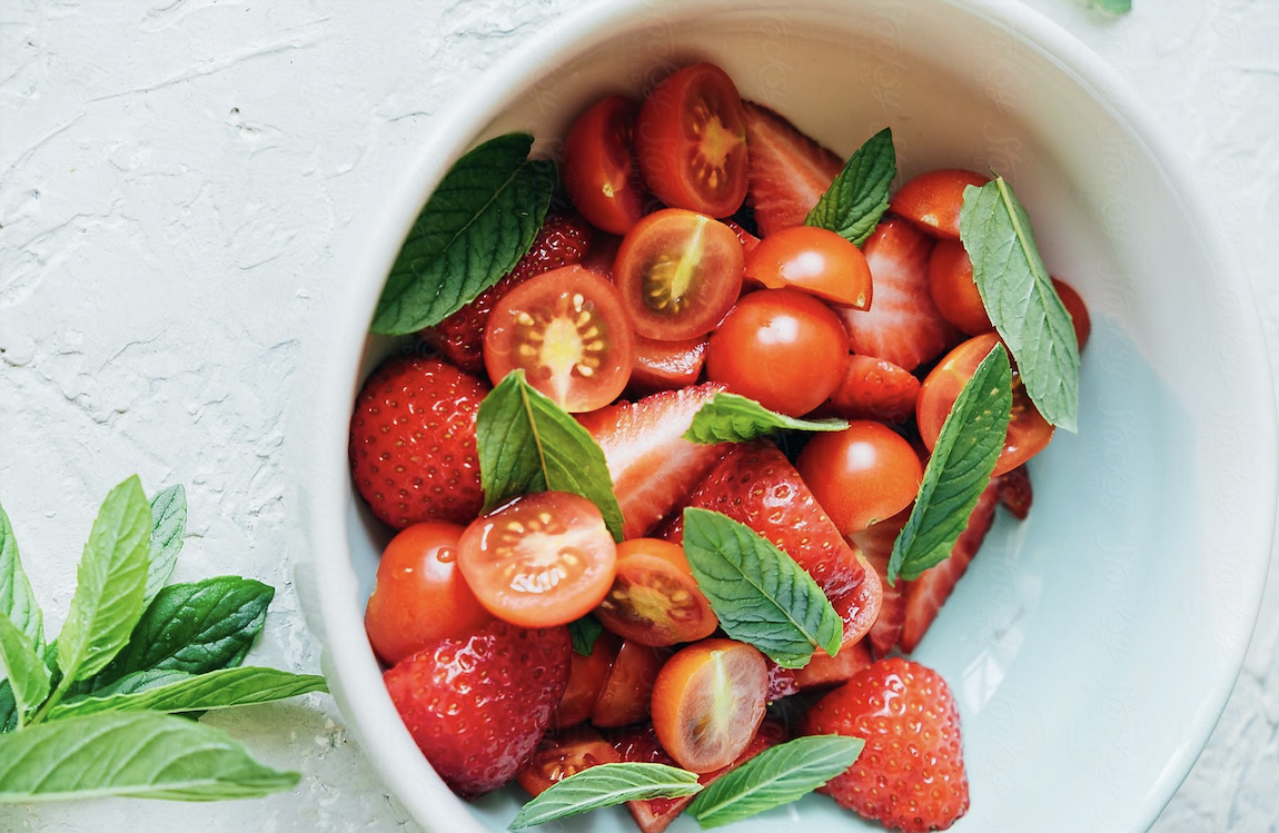 A white bowl filled with chopped strawberries, cherry tomatoes, and fresh mint leaves on a textured white background. Mint leaves are also arranged beside the bowl. Perfect for celebrating your latest plant clearance sale haul!