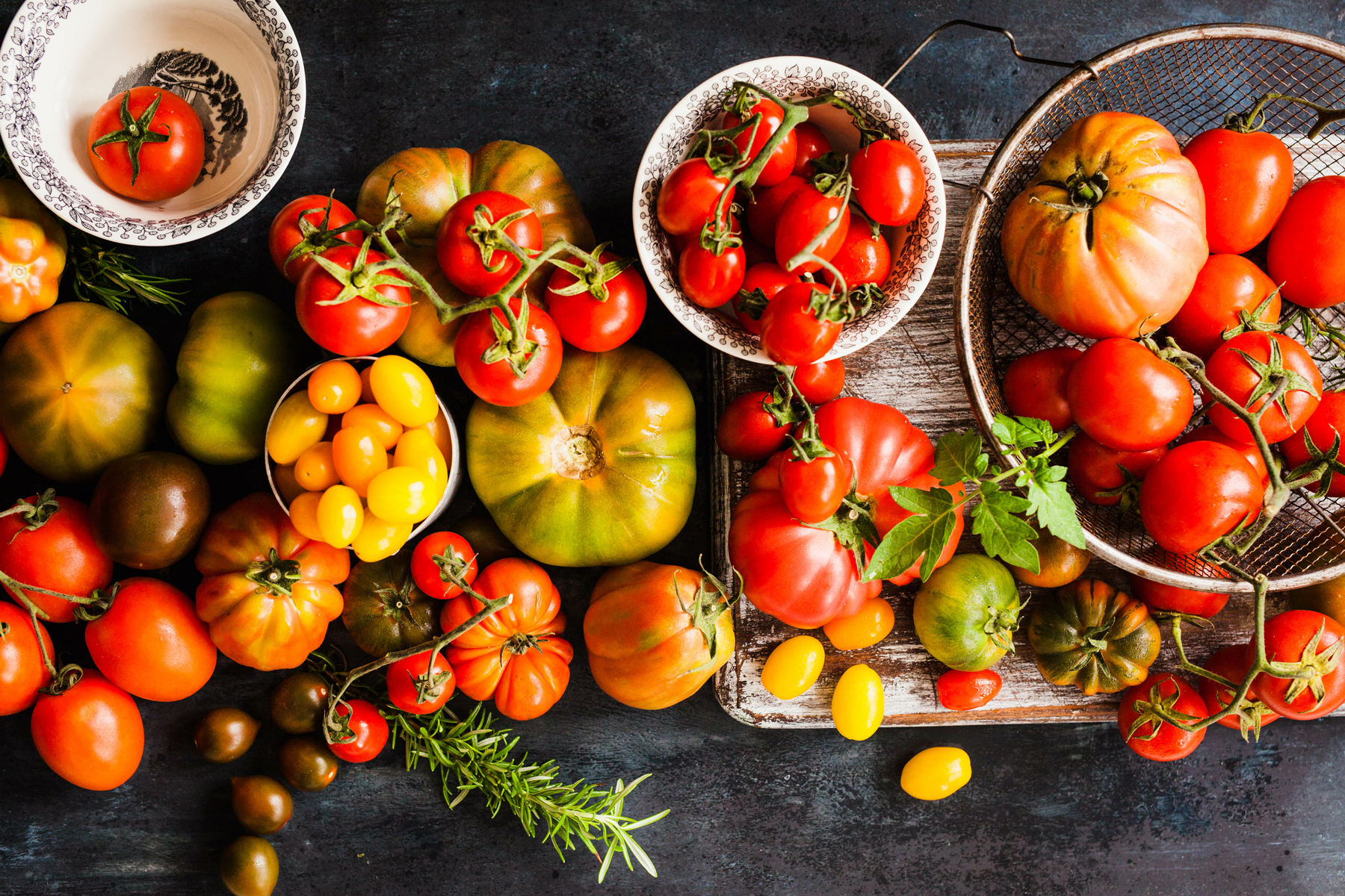 A variety of tomatoes in different shapes and colors, some in bowls and others on a table, with a sprig of rosemary nearby, showcasing the solution to many garden design problems.