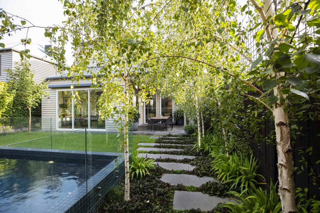 A Hamptons garden features a backyard with a small pool, stone pathway, and lush greenery. A modern house stands in the background, complete with large windows that invite natural light to dance across the serene landscape.