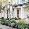 Front view of a house with a manicured Hamptons garden featuring shrubs, grasses, and decorative pots. The house boasts a white exterior, large windows, and a covered porch with elegant columns.