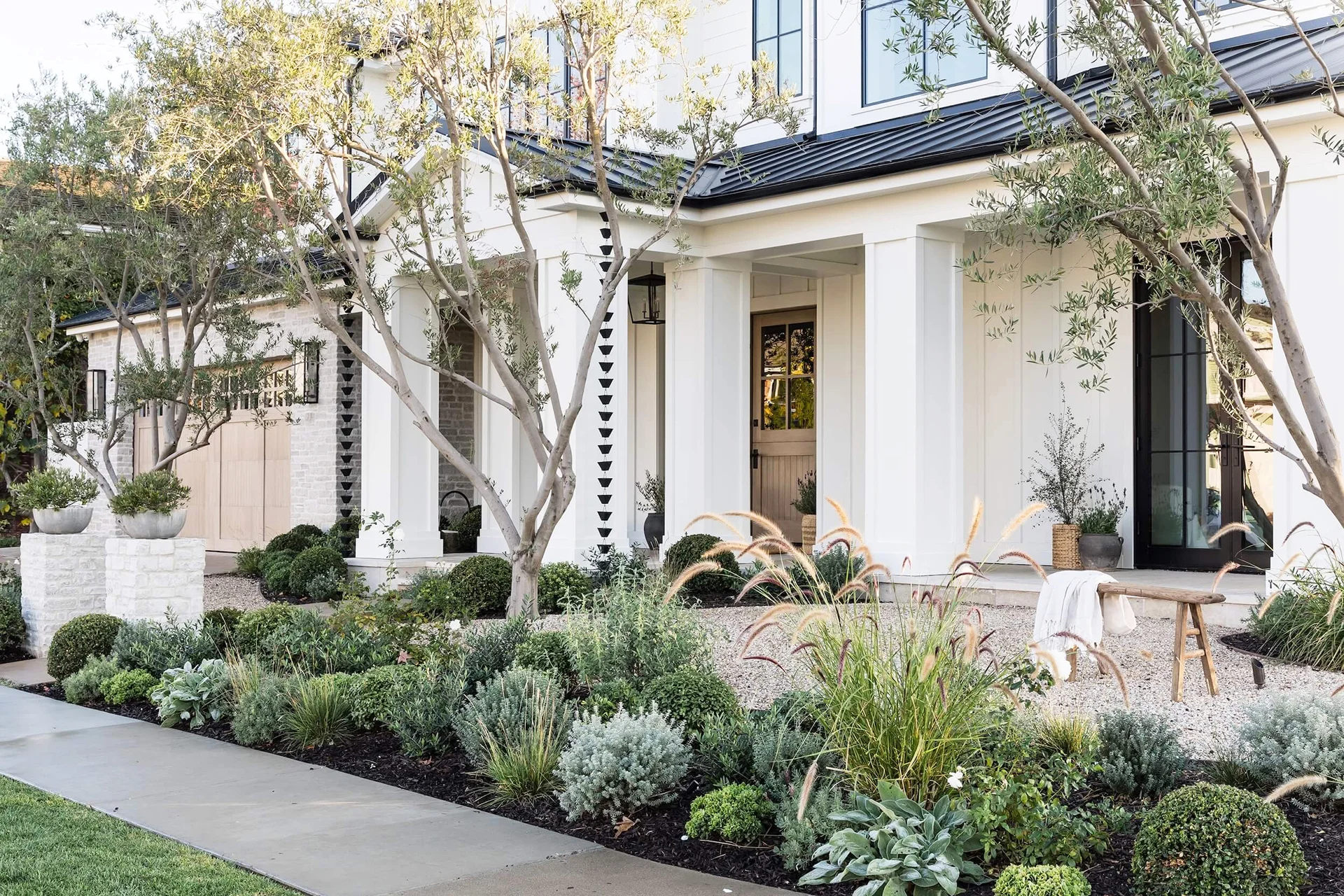 Front view of a house with a manicured Hamptons garden featuring shrubs, grasses, and decorative pots. The house boasts a white exterior, large windows, and a covered porch with elegant columns.