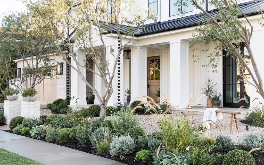 A modern house with a porch overlooks its Hamptons-inspired garden, where neatly landscaped shrubs, ornamental grasses, and potted plants create a serene front yard.