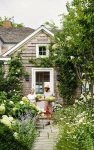 Two people standing in a lush garden, holding hydrangeas, in front of a shingled house with a white door. The garden path is lined with various flowering plants.