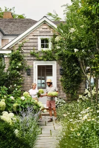 Two people stand on a garden path in front of a charming Hamptons house, holding hydrangeas. The home features classic shingles and a white door, surrounded by lush greenery and vibrant flowers, capturing the essence of a serene Hamptons garden.