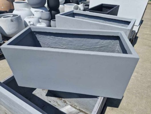 The sunlit outdoor surface features multiple rectangular gray concrete planters, such as the GardenLite Trough Cement M model with dimensions of 80x37x37cm.