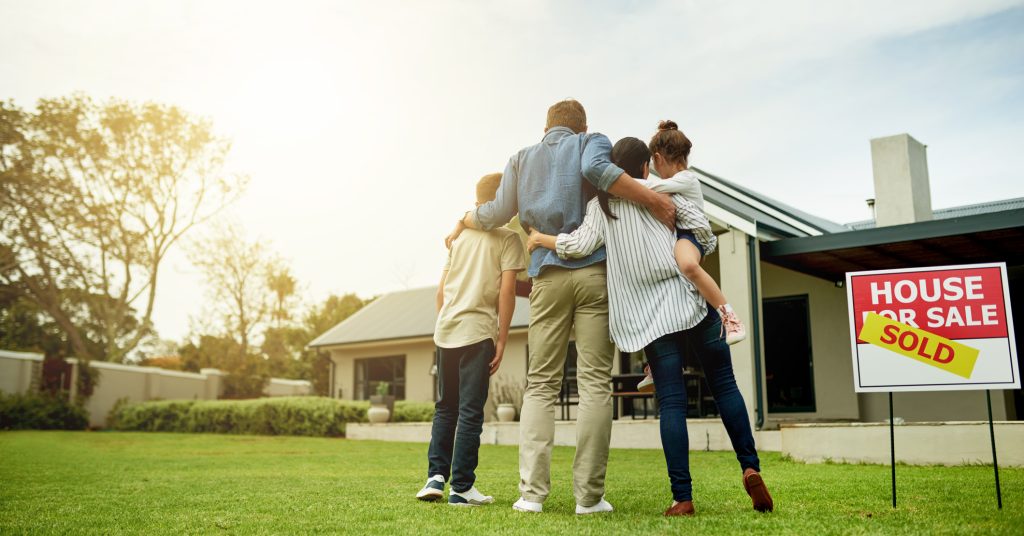 A family stands joyously in front of their new home, where alongside the "House for Sale" sign marked "Sold," a row of Christmas trees adds a festive touch to their fresh start.