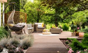 A wooden deck with wicker furniture, cushions, potted plants, and greenery, surrounded by lush trees and bushes.