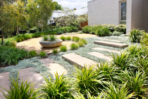 Modern garden with stepping stones, a round planter, and greenery surrounding a gravel path next to a white building.