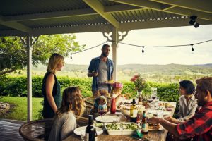 A group of people enjoy a meal on a covered porch overlooking a scenic landscape. The table is set with food and drinks.