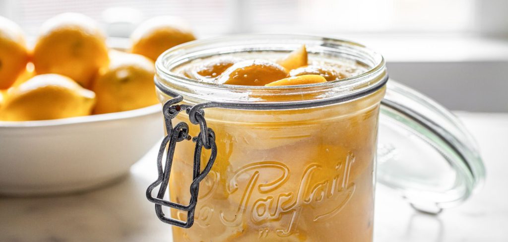 A glass jar filled with preserved lemons sits on a marble countertop, reminiscent of ornaments, with a bowl of fresh lemons in the background. The scene is softly lit by natural light, evoking the charm of Christmas trees.