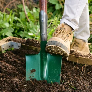 A person wearing brown boots uses a green shovel to dig soil in a garden.