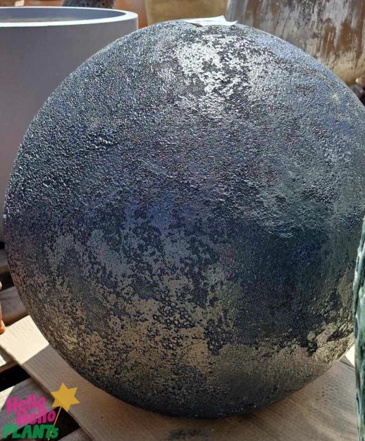 A large, textured, spherical object with a dark, speckled surface reminiscent of Iridami Relief sits on a wooden pallet. The object is the Sorrento Decorative Ball in black, measuring 50x50cm.