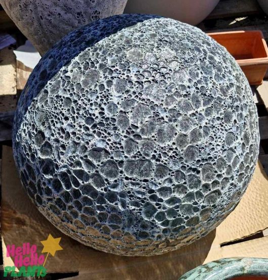 The Sorrento Decorative Ball Grey 50x50cm, with its large textured spherical form and Iridami relief, resembles Dewi Sri's serene beauty and stands on a wooden pallet among other garden items, its crater-like surface creating an enchanting display.