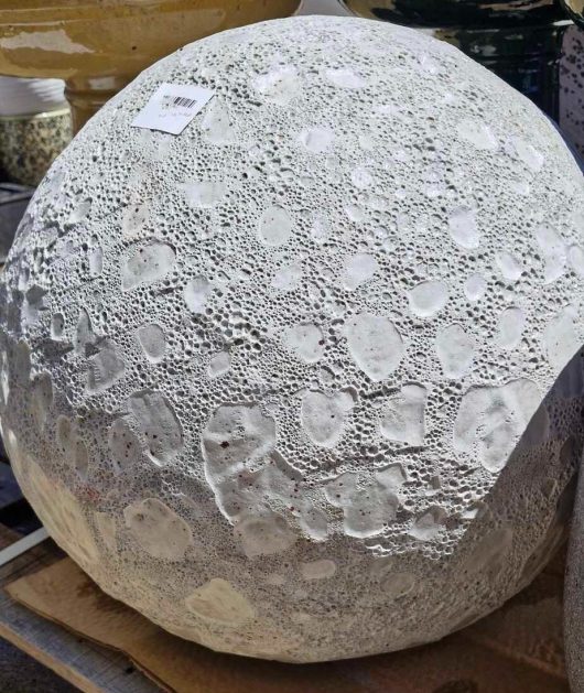 Close-up of a large, textured stone sphere with a rough, crater-like surface resembling the Sorrento 'Sadiki Urn Antique' M 40x36cm. A small price tag adorns this intriguing outdoor display piece.