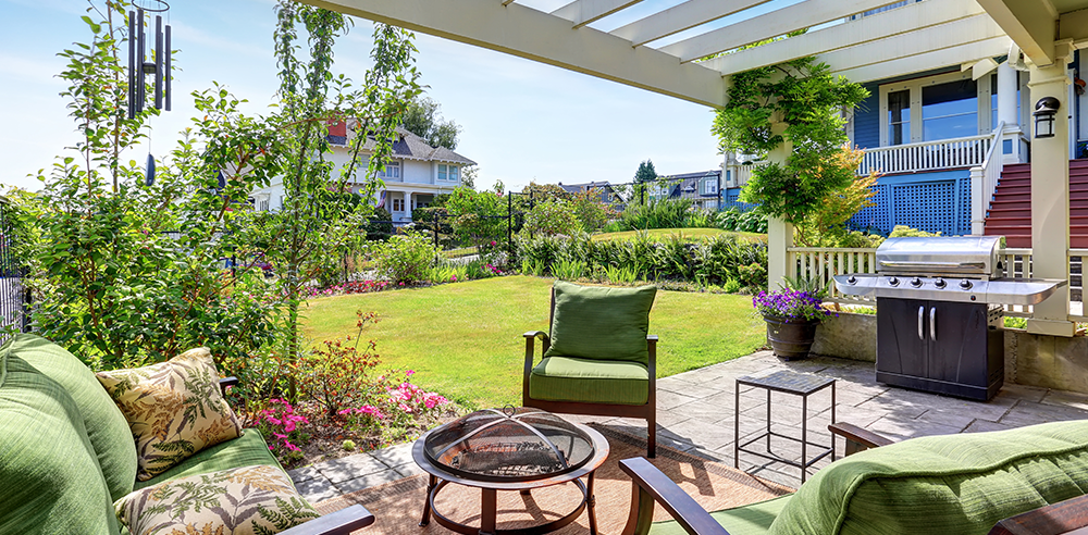 Enjoy the many benefits of gardening in this serene outdoor patio, featuring green cushioned seating around a round table and a gas grill. A lush garden and lawn create the perfect backdrop under a clear sky.