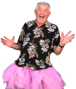 Older man wearing a floral shirt and pink tutu, smiling with arms raised against a plain background.