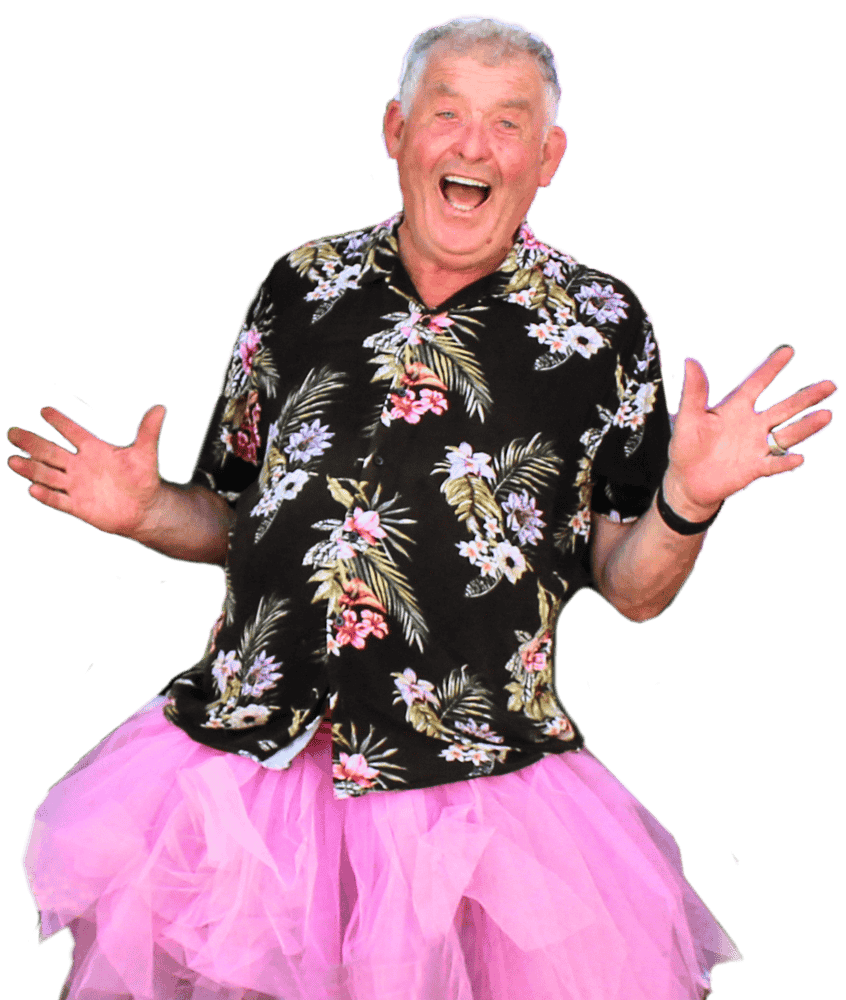 Older man wearing a floral shirt and pink tutu, smiling with arms raised against a plain background.