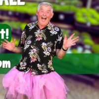 An older man in a floral shirt and pink tutu smiles widely with arms up, as if celebrating life at a vibrant plant sale, standing against a blurred, colorful background.