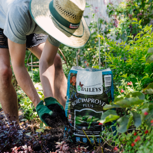 Person wearing a straw hat and gloves, spreading soil from a bag labeled "Baileys Soil Improver Plus" in a garden.