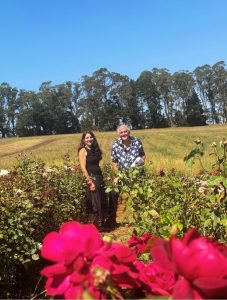 Two people stand in a field surrounded by blooming flowers and lush greenery, with the sweet smell of roses wafting through the air. Trees frame the scene under a clear blue sky, completing this serene setting.