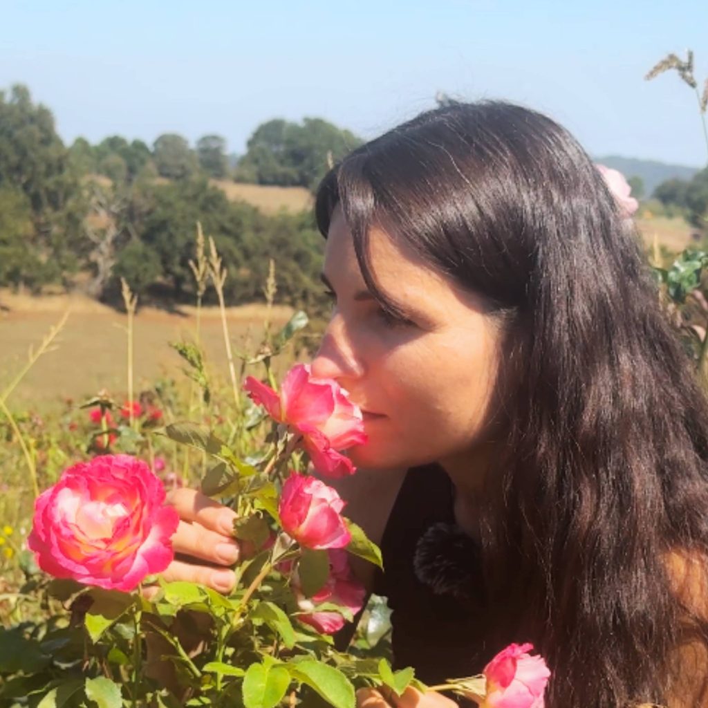 In a garden setting, with fields and trees stretching into the background, a person with long dark hair savors the rose smell of pink blooms.